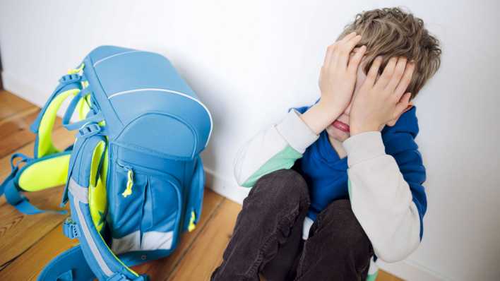 Ein Junge sitzt traurig in seinem Kinderzimmer neben seinem Schulranzen und versteckt sein Gesicht hinter seinen Haenden.