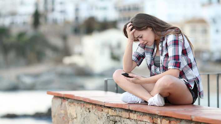 Ein Mädchen sitzt auf einer Mauer und schaut verzweifelt auf ihr Handy.