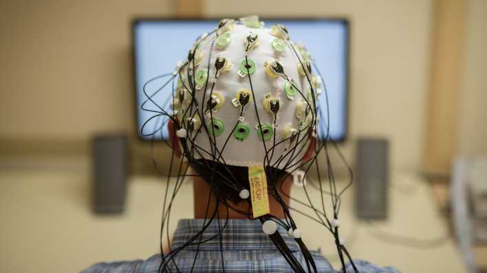 Ein Mann sitzt mit Elektroden auf dem Kopf vor einem Bildschirm.