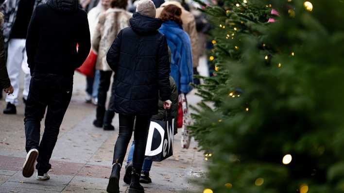 Eine Frau trägt in einer belebten Fußgängerzone eine Einkaufstüte aus Papier und läuft gerade an einem Nadelbaum vorbei, der mit Lichtern geschmückt ist.