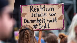 Bei einer Demonstration hält eine Teilnehmerin ein Schild mit der Aufschrift "Reichtum schützt vor Dummheit nicht #Sylt" hoch.
