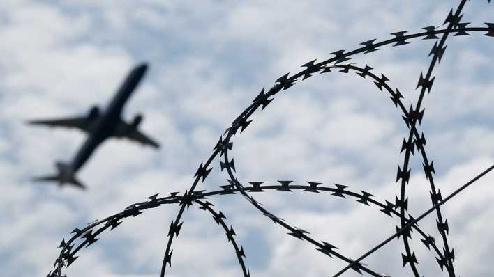 Ein Flugzeug hebt am Flughafen BER hinter einem Zaun mit Natodraht ab.