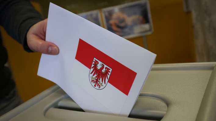 Ein Stimmzettel mit dem Wappen von Brandenburg wird in eine Urne geworfen.