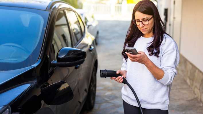 Eine Frau steht mit Ladekappel und Smartphone neben einem Elektroauto (Bild: picture alliance / Westend61 | William Perugini)