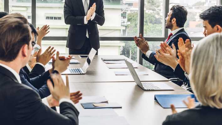 Bei einem Meeting applaudieren viele Mitarbeiter einem Geschäftsmann.