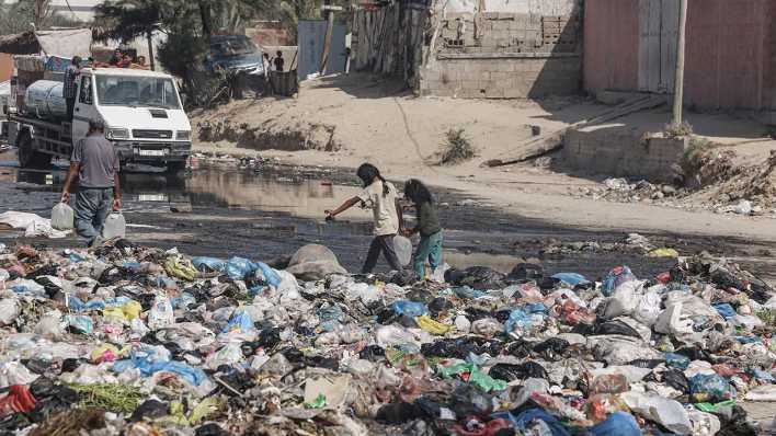 Zwei Mädchen tragen in Dair El balah Wasserkanister über eine mit Mülltüten verunreinigte Straße.