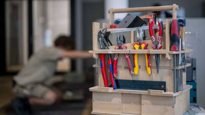 Ein Werkzeugkasten steht auf dem Tisch, während im Hintergrund ein Handwerker ein Möbelstück zusammenbaut.