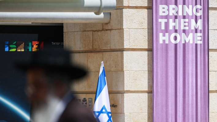 Mit Blick auf die von der Hamas verschleppten israelischen Geiseln steht "Bring them home" (Bringt sie heim) auf einem Transparent in Jerusalem, im Vordergrund geht ein orthodoxer Jude vorbei. (Bild: picture alliance/dpa/Hannes P. Albert)