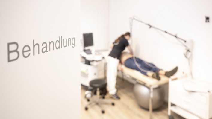 An einer Tür steht "Behandlung", im Hintergrund sieht man einen Patienten auf einer Liege.