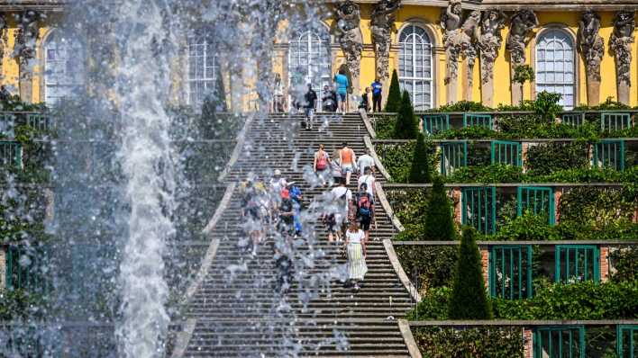 Hinter der Großen Fontäne sind bei sommerlichem Wetter und hohen Temperaturen zahlreiche Touristen am Schloss Sanssouci in Potsdam unterwegs.