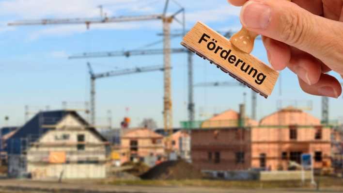 Über ein Neubaugebiet mit Wohnungen und Häusern im Rohbau wird ein Holzstempel gehalten mit dem Aufdruck: Förderung