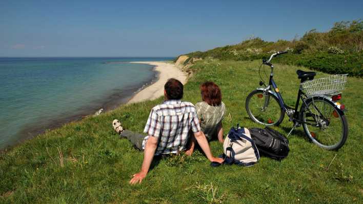 Ein Paar sitzt auf einem Rasen und schaut aus der Ferne auf Strand und Meer (Bild: picture alliance | CHROMORANGE / Dieter Möbus)