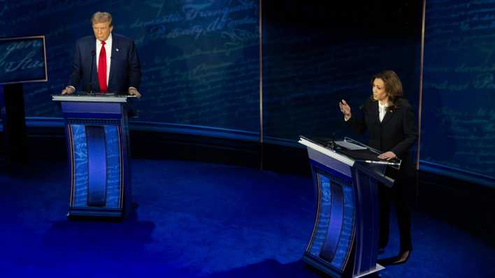 Donald Trump und Kamala Harris stehen während des Fernsehduells in einem in blau gehaltenen Fernsehstudio.