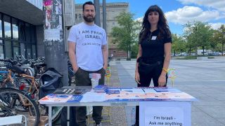 Shay Dashevsky steht an einem Stand vor einer Universität und trägt ein T-Shirt mit der Aufschrift "I am from Israel - aks me anything".