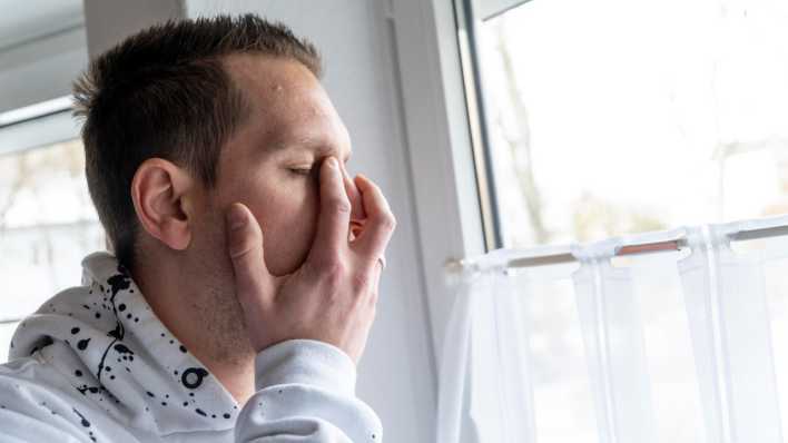 Ein junger Mann steht traurig und verzweifelt an einem Fenster, er hält sich das Gesicht vor Schmerz.