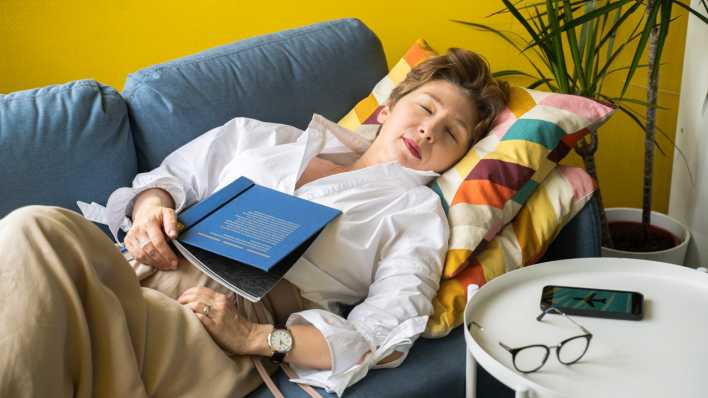 Eine Frau ist mit einem Buch auf dem Bauch auf einem Sofa eingeschlafen.