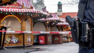 Eine Schusswaffe am Gürtel eines Polizeibeamten, im Hintergrund ist ein Absperrband und der Magdeburger Weihnachtsmarkt zu sehen.