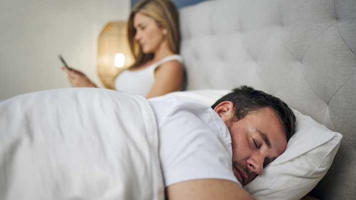 Ein Mann schläft im Doppelbett - seine Frau sitzt neben ihm und hat das Handy in der Hand.