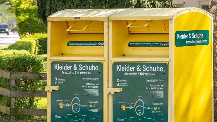 Zwei gelbe Altkleidercontainer mit der Aufschrift "Kleider & Schuhe Accessoires & Heimtextilien" auf einem Parkplatz.