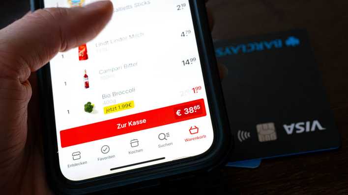 Eine Kreditkarte liegt neben einem Smartphone, auf dem eine App für den Einkauf von Lebensmitteln geöffnet ist (Bild: picture alliance/dpa/Sina Schuldt)
