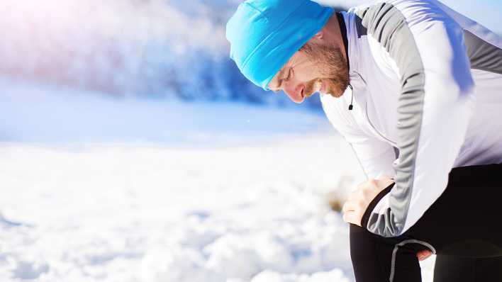 Ein Mann in Sportkleidung hält sich in einer Schneelandschaft das Knie.