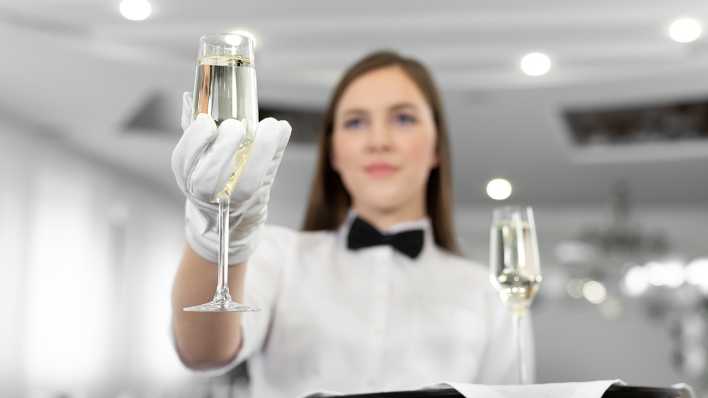 Eine Frau im weißen Hemd mit schwarzer Fliege serviert mit weißen Handschuhen ein Glas Champagner.