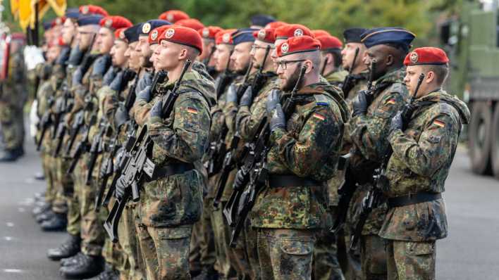 Eine Gruppe von Bundeswehrsoldaten in Tarnuniform und mit roten Baretten steht während einer Zeremonie mit Gewehren im Anschlag in einer Kaserne.